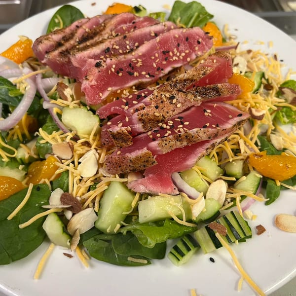 Seared tuna salad with spinach, cucumbers, oranges, cheese, and almonds on a white plate.