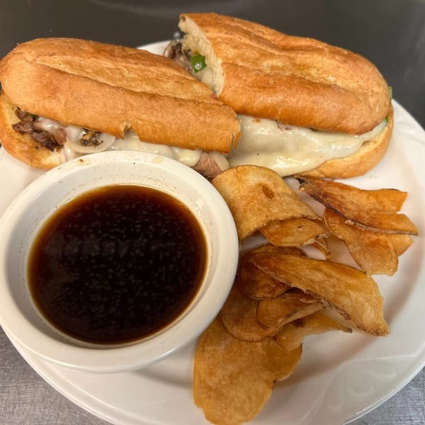 A ShoeBooties Cafe Philly cheesesteak sandwich with a side of au jus and crispy chips.